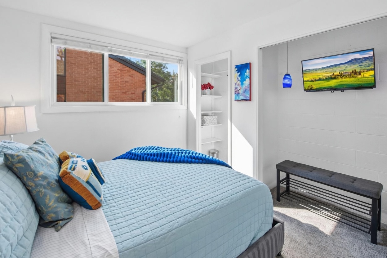 This bright bedroom features a comfortable bed dressed in a light blue quilt with decorative accent pillows. To the side, a built-in shelving unit provides storage, while a wall-mounted television is positioned above a dark, tufted bench. The room is illuminated by natural light from a large window and a decorative blue pendant light, creating a clean and inviting atmosphere.
