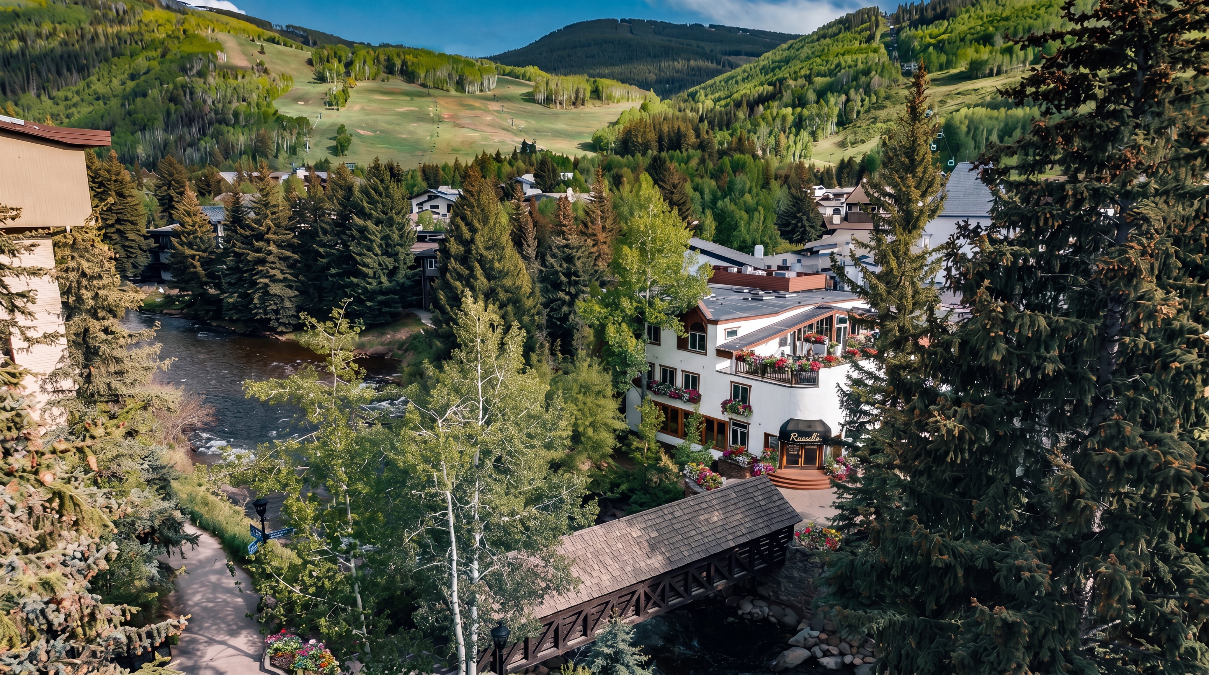 This elevated aerial perspective captures a scenic mountain resort setting, featuring a winding river flowing past a charming covered wooden bridge. A multi-story building with a distinctive white facade and dark roof sits nestled among lush green trees and dense forest. In the background, a vast, grassy ski slope stretches up the mountainside, emphasizing the property's prime location for outdoor recreation.