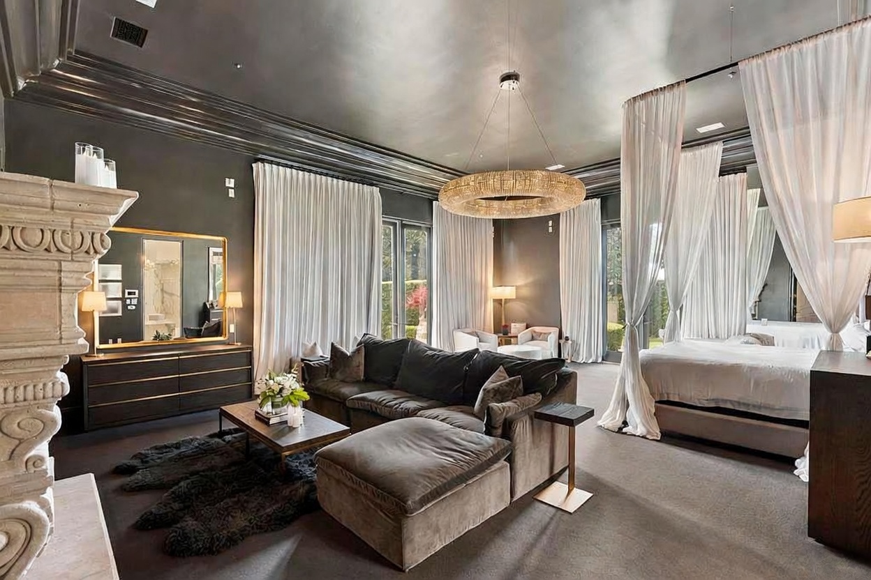 This is a luxurious primary bedroom featuring a four-poster bed draped with sheer white curtains, a comfortable seating area with a large sectional sofa and ottoman, and a statement chandelier. The room is decorated in a sophisticated color palette of grays and browns, with a large mirror and fireplace adding to the elegant ambiance. The room is spacious and well-lit, creating a relaxing and inviting atmosphere.