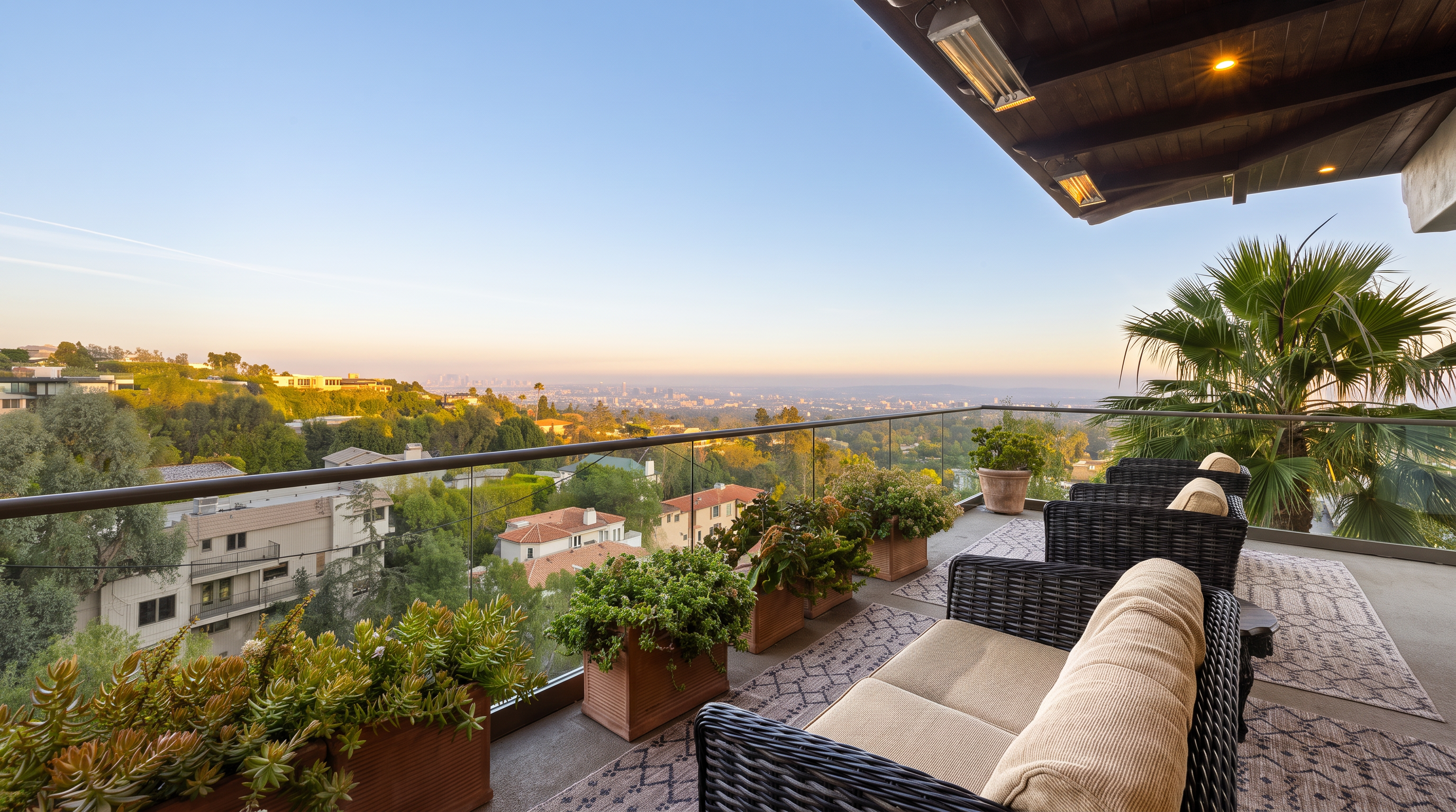 This elevated balcony offers a panoramic view of the city skyline at sunset, framed by a sleek glass railing. The space is furnished with comfortable wicker-style seating topped with neutral cushions and accented by several large planters filled with lush greenery. A covered ceiling with recessed lighting and mounted heaters provides a cozy, sheltered atmosphere for outdoor relaxation.
