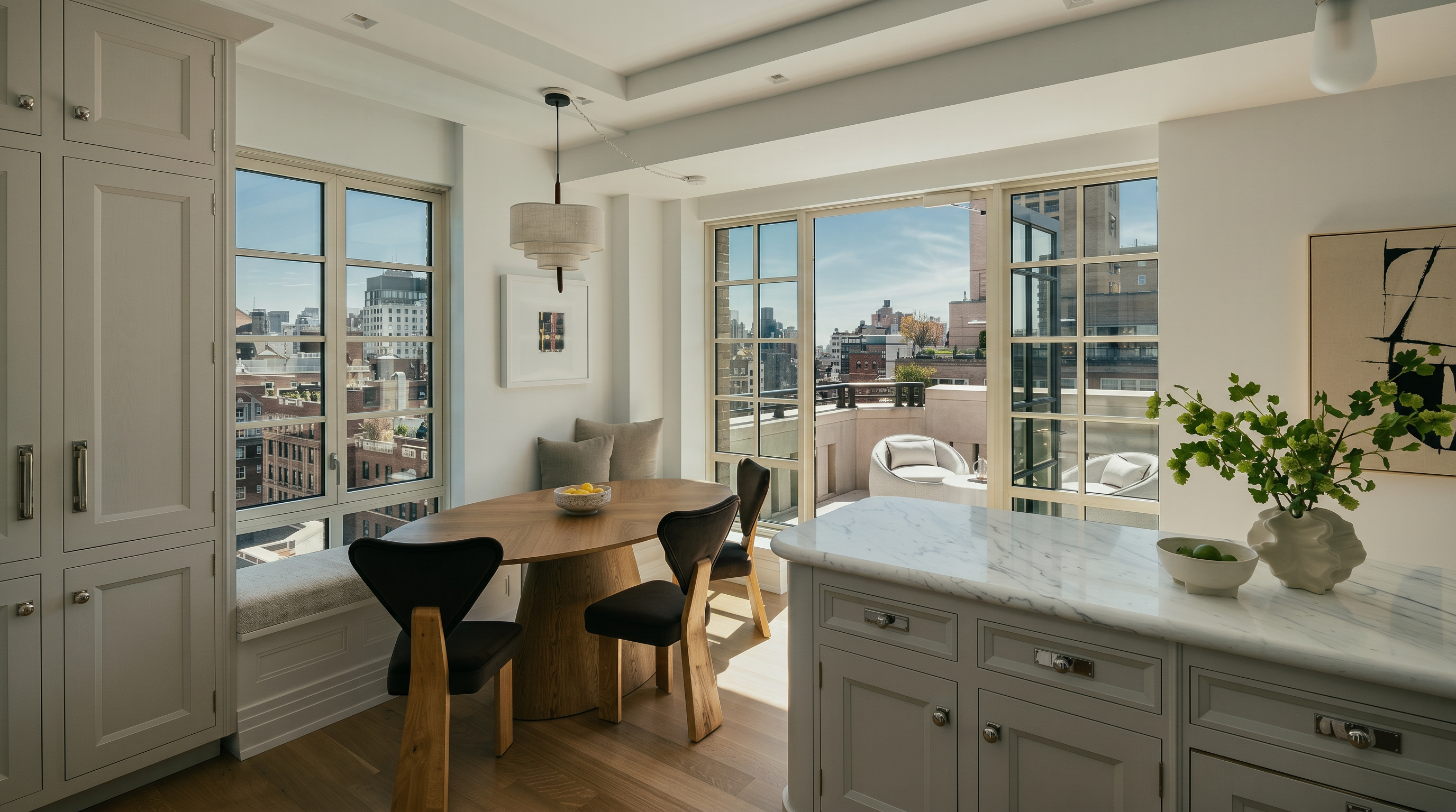 This bright and airy kitchen features a central island with a stone countertop and light-toned cabinetry, seamlessly transitioning into a cozy dining nook. The space is defined by large windows that provide abundant natural light and views of the city skyline and an adjacent outdoor terrace. The combination of light wood flooring, elegant pendant lighting, and a neutral color palette creates a sophisticated and inviting atmosphere.