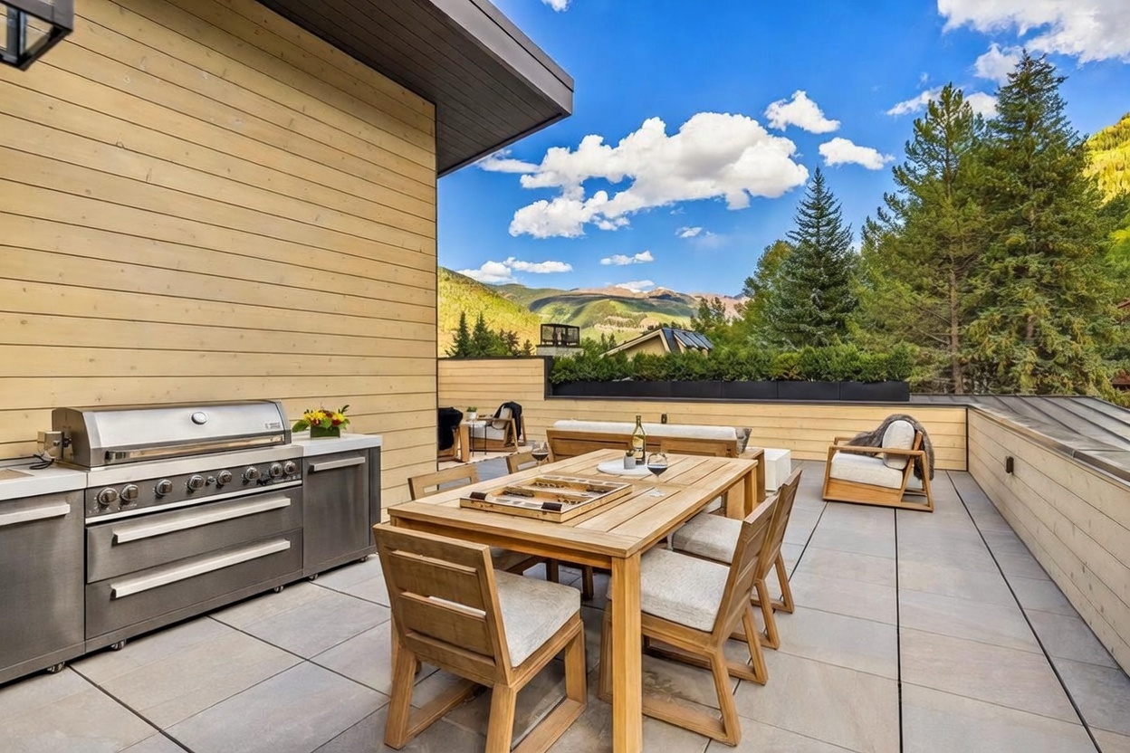 This expansive rooftop deck features a modern outdoor kitchen with a stainless steel grill and a light wood dining set arranged for al fresco entertaining. The space is finished with large-format stone tiles and surrounded by light wood siding, offering panoramic views of the surrounding mountains and pine trees. A comfortable lounge area with plush seating completes this sophisticated outdoor living space, perfect for enjoying the scenic landscape.
