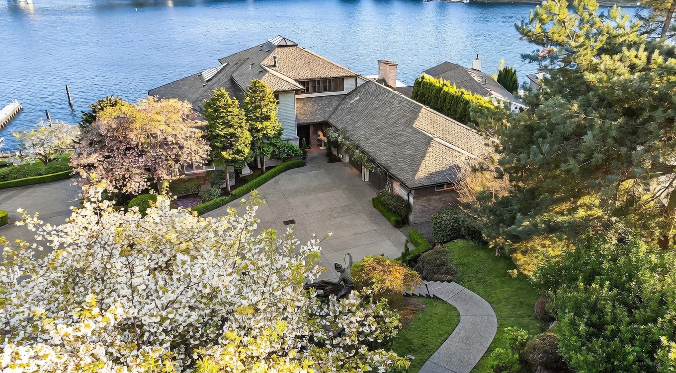 This high-angle aerial perspective captures a sprawling waterfront estate nestled among lush, mature trees and vibrant spring foliage. The home features a complex, multi-gabled roofline and a spacious concrete motor court leading to a multi-car garage, with a winding stone pathway guiding visitors toward the entrance. The property enjoys a serene setting directly adjacent to a calm body of water, offering a picturesque blend of architectural elegance and natural beauty.