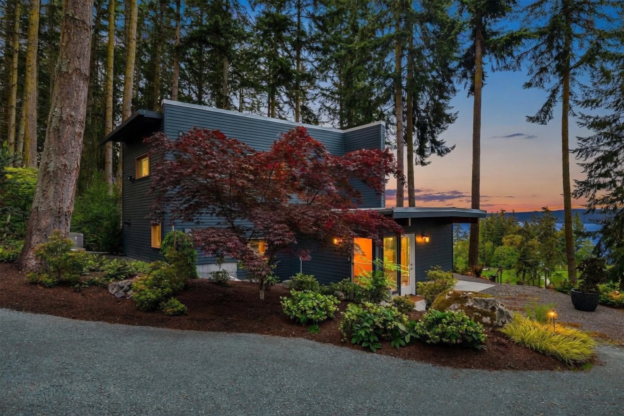 This modern, dark-sided home is nestled within a lush, wooded landscape, featuring a striking architectural design with clean lines and large windows. A prominent Japanese maple with deep red foliage stands in the foreground, contrasting beautifully against the dark exterior and the twilight sky. The gravel driveway leads toward the home's entrance, which is warmly illuminated, creating an inviting atmosphere against the backdrop of a distant water view.