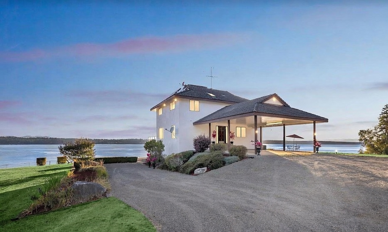 This waterfront property features a white-sided home with a prominent covered entryway and a dark-tiled roof. A long, gravel driveway leads up to the house, which is set against a scenic backdrop of a calm body of water and distant hills under a soft, twilight sky. The landscaping includes manicured green lawns and natural shrubbery, creating a serene and inviting atmosphere.