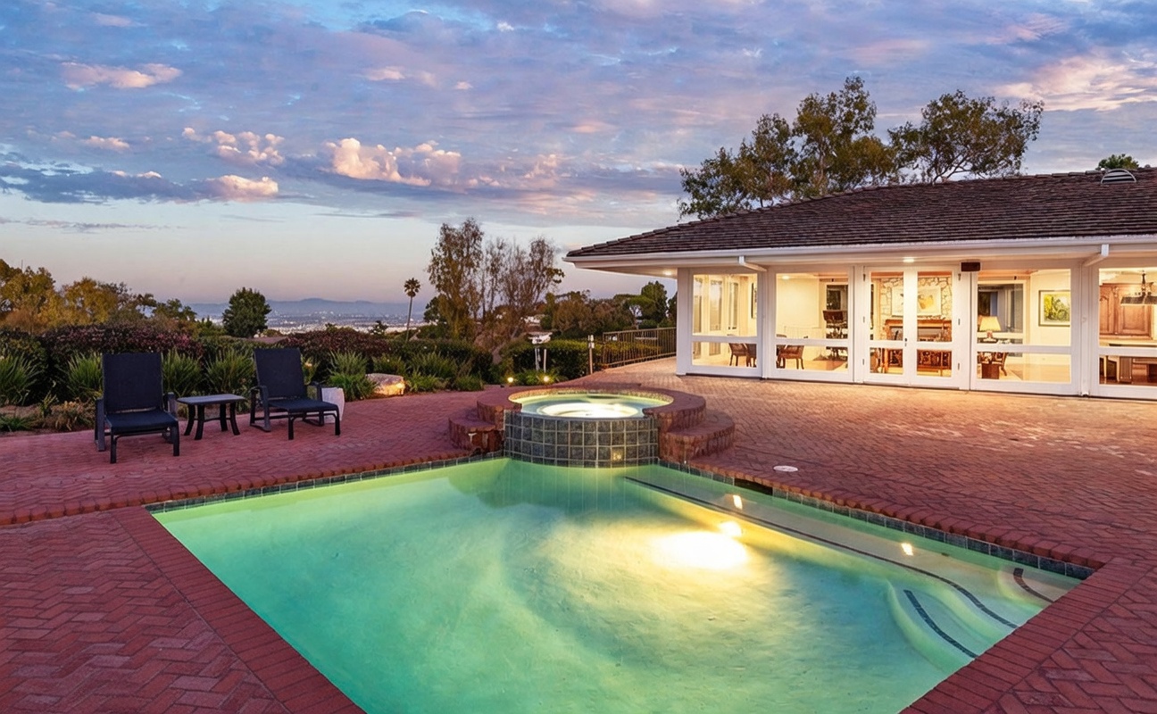 This rear exterior view showcases a spacious brick-paved patio featuring a built-in circular spa that spills into a swimming pool. The home features large glass doors that open to the outdoor living area, providing a seamless transition between indoor and outdoor spaces. The scene is set against a scenic backdrop of rolling hills and a distant cityscape under a vibrant sunset sky.
