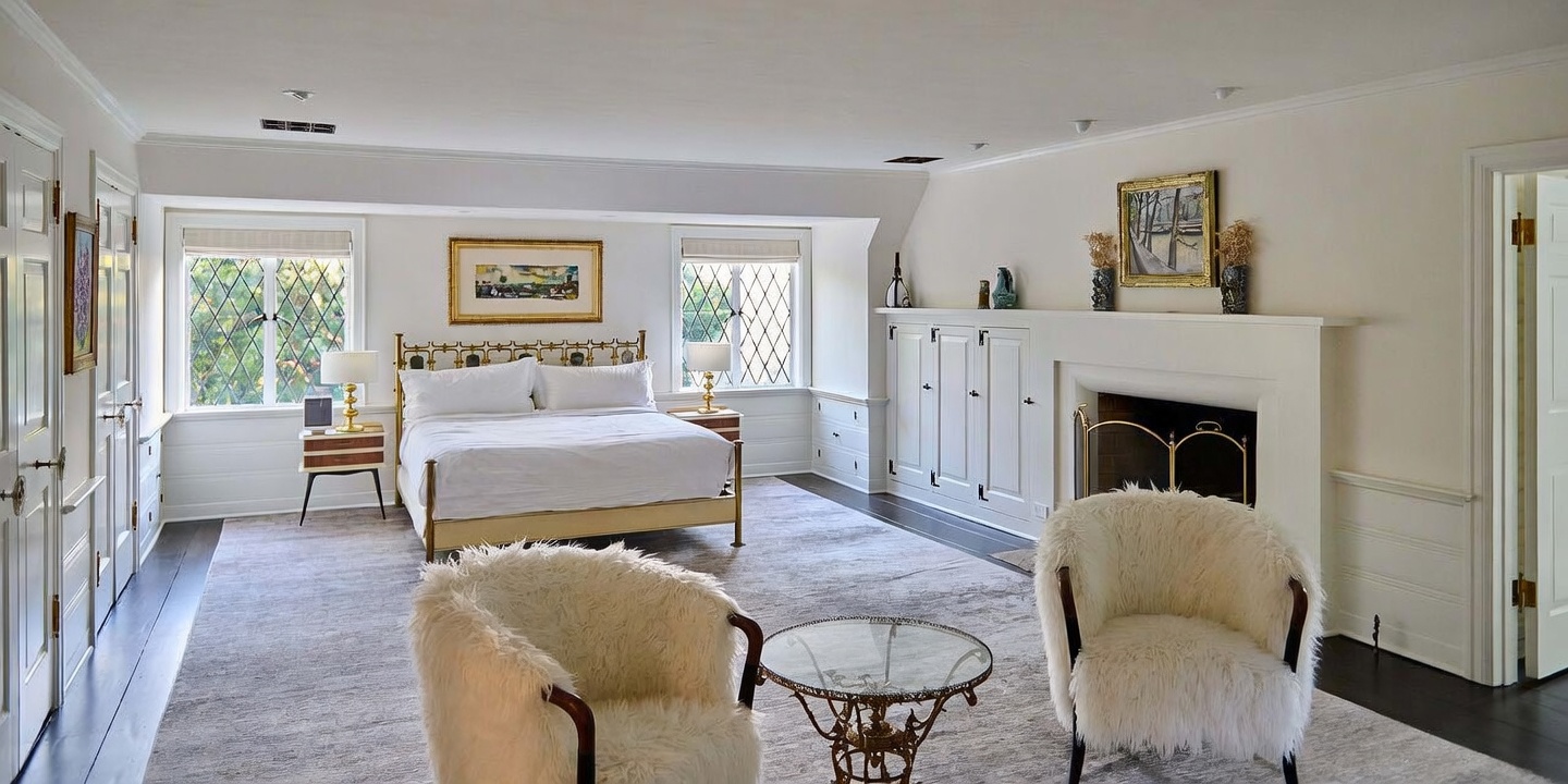 This spacious primary bedroom features a classic, elegant design with a large bed centered between two windows and a cozy seating area in the foreground. A fireplace with a white mantel and built-in cabinetry adds architectural interest, while the neutral color palette and plush carpeting create a serene and inviting atmosphere. The room is well-lit with natural light, offering a sophisticated and comfortable retreat.