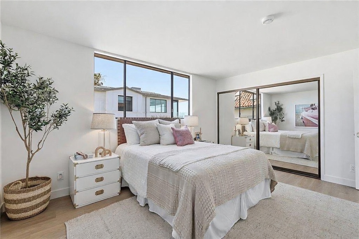 This bright and airy bedroom features a queen-sized bed with neutral-toned bedding and a textured headboard, centered in front of a large window that allows for ample natural light. To the left, a white three-drawer nightstand holds a lamp, while a large mirrored closet door on the right reflects the room, enhancing the sense of space. The room is finished with light wood-toned flooring and a neutral area rug, creating a clean and inviting atmosphere.
