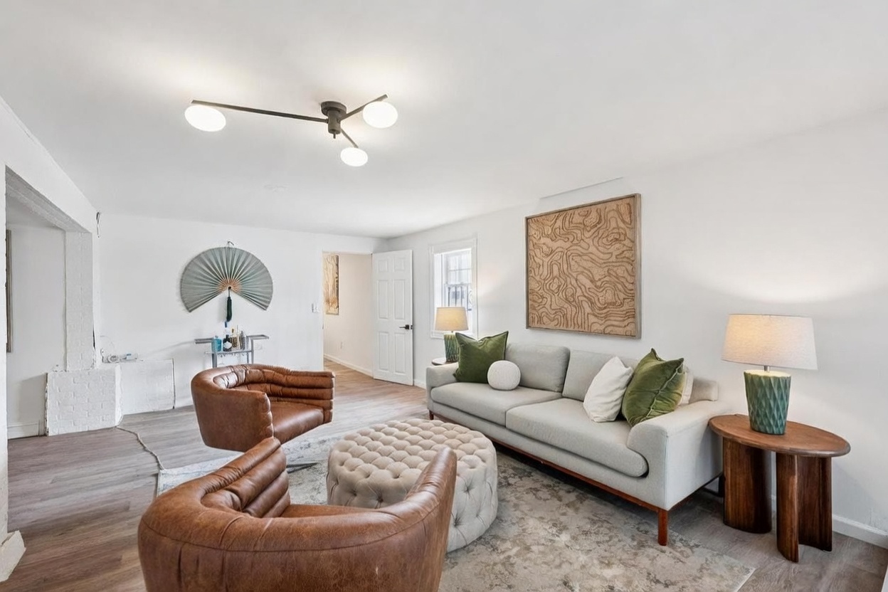 This bright and inviting living room features a neutral color palette with warm leather accent chairs and a tufted ottoman. A light gray sofa is adorned with green and white throw pillows, positioned beneath a large, textured wall art piece. The space is illuminated by a modern multi-arm ceiling fixture and a table lamp, creating a comfortable and stylish atmosphere.