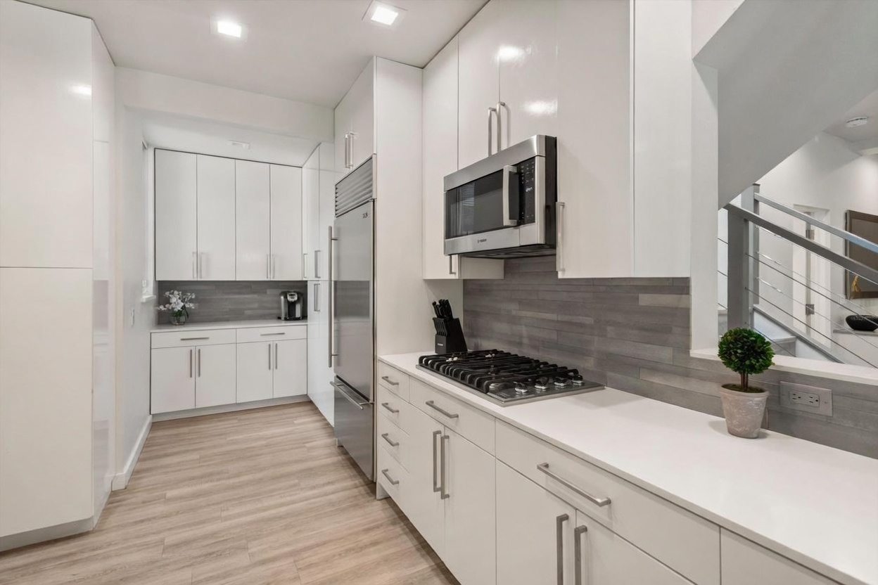 This modern, galley-style kitchen features sleek white cabinetry, stainless steel appliances, and a light-toned wood-look floor. A grey horizontal tile backsplash adds texture to the space, which is illuminated by recessed ceiling lights and includes a gas cooktop and built-in microwave. The perspective captures the efficient layout, showing the transition toward a staircase with a metal railing on the right side.