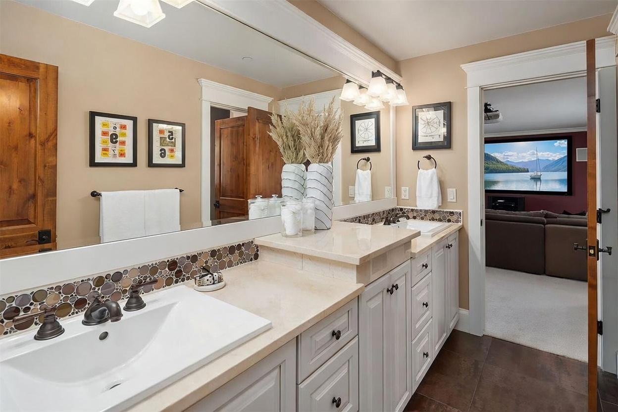This bathroom features a dual-sink vanity with light-colored cabinetry and a light-toned stone countertop, accented by a decorative mosaic tile backsplash. A large mirror spans the wall above the vanity, reflecting the room and the adjacent doorway that leads into a media room with a large screen. The space is well-lit by a multi-bulb vanity light fixture and includes framed wall art, creating a polished and functional aesthetic.