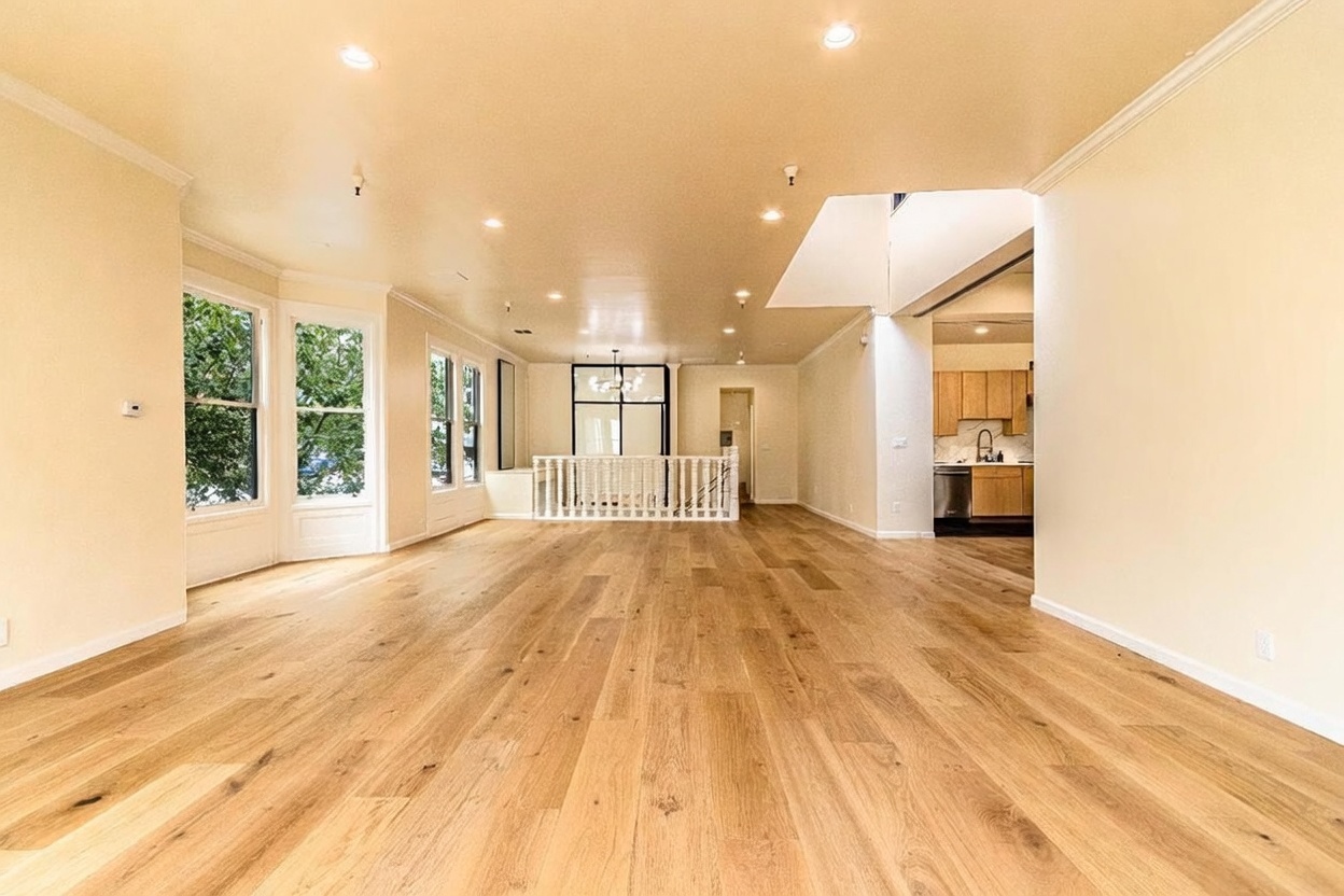 This spacious, open-concept living area features light wood flooring and neutral walls, creating a bright and airy atmosphere. The room is illuminated by numerous recessed ceiling lights and includes a white railing that overlooks a lower level, with a glimpse of a kitchen visible in the background. Large windows on the left side allow for natural light, contributing to the clean and modern aesthetic of the home.