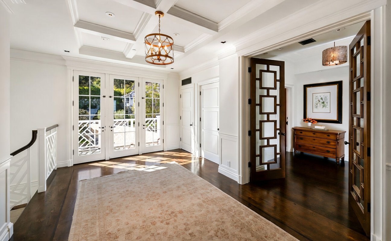 This elegant foyer features dark hardwood flooring, a coffered ceiling with a decorative chandelier, and a large, neutral-toned area rug. A set of glass-paned double doors leads to the exterior, while an ornate, dark-wood decorative door opens into an adjacent room containing a wooden chest and framed artwork. The space is bright and airy, showcasing a sophisticated architectural design with crisp white trim and wainscoting.