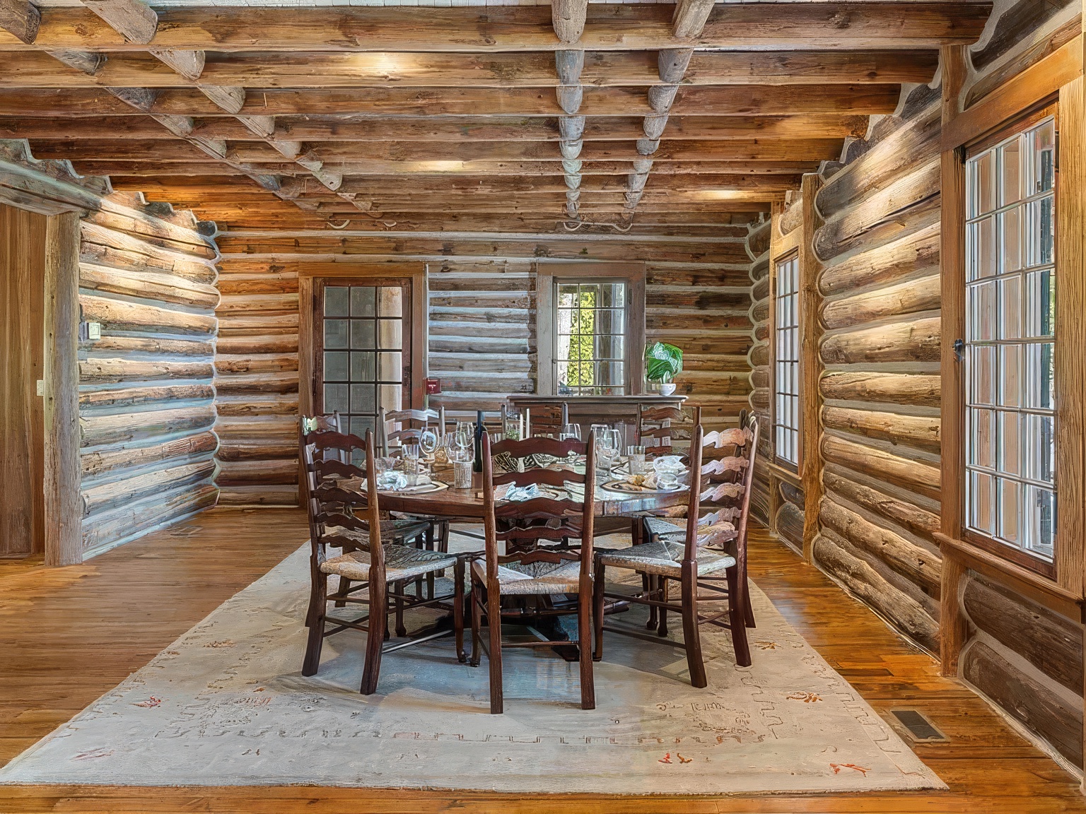 This rustic dining room features exposed log walls and a beamed ceiling, creating a warm and authentic cabin atmosphere. A circular wooden table is surrounded by six spindle-back chairs, all set atop a large, light-colored area rug that anchors the space. The room is illuminated by natural light streaming through multiple windows, including a large window on the right and a glass-paned door in the background.