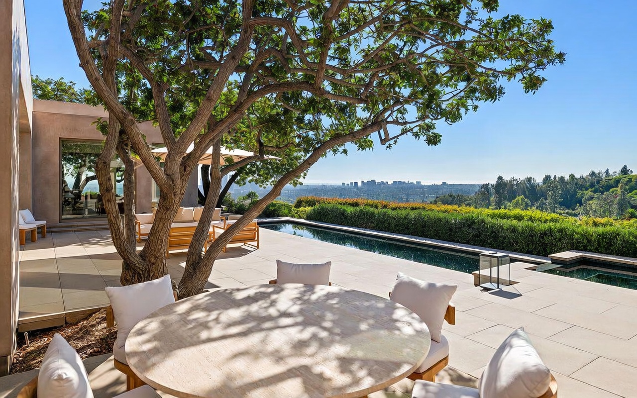 This luxurious outdoor patio space features a stone-topped dining table and comfortable lounge seating, all shaded by a large, mature tree. The area seamlessly transitions to a sleek, rectangular infinity-edge pool that overlooks a sprawling cityscape in the distance. The overall aesthetic is one of sophisticated, modern relaxation, perfectly designed for high-end outdoor entertaining.