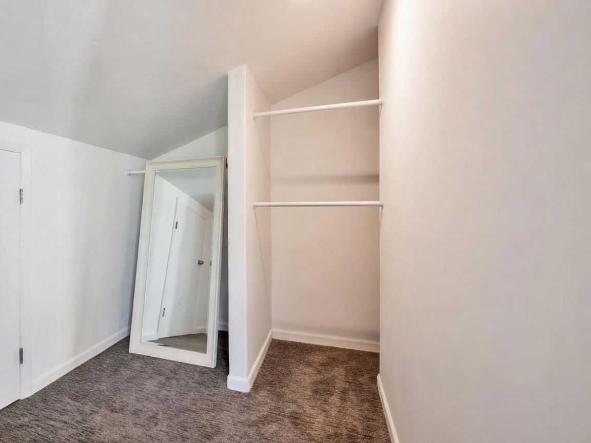 This walk-in closet space features a slanted ceiling, clean white walls, and dark gray wall-to-wall carpeting. It includes a floor-standing white-framed mirror and a dedicated recessed area with two white clothing rods for storage. The overall design is functional and minimalist, creating a neat storage solution.