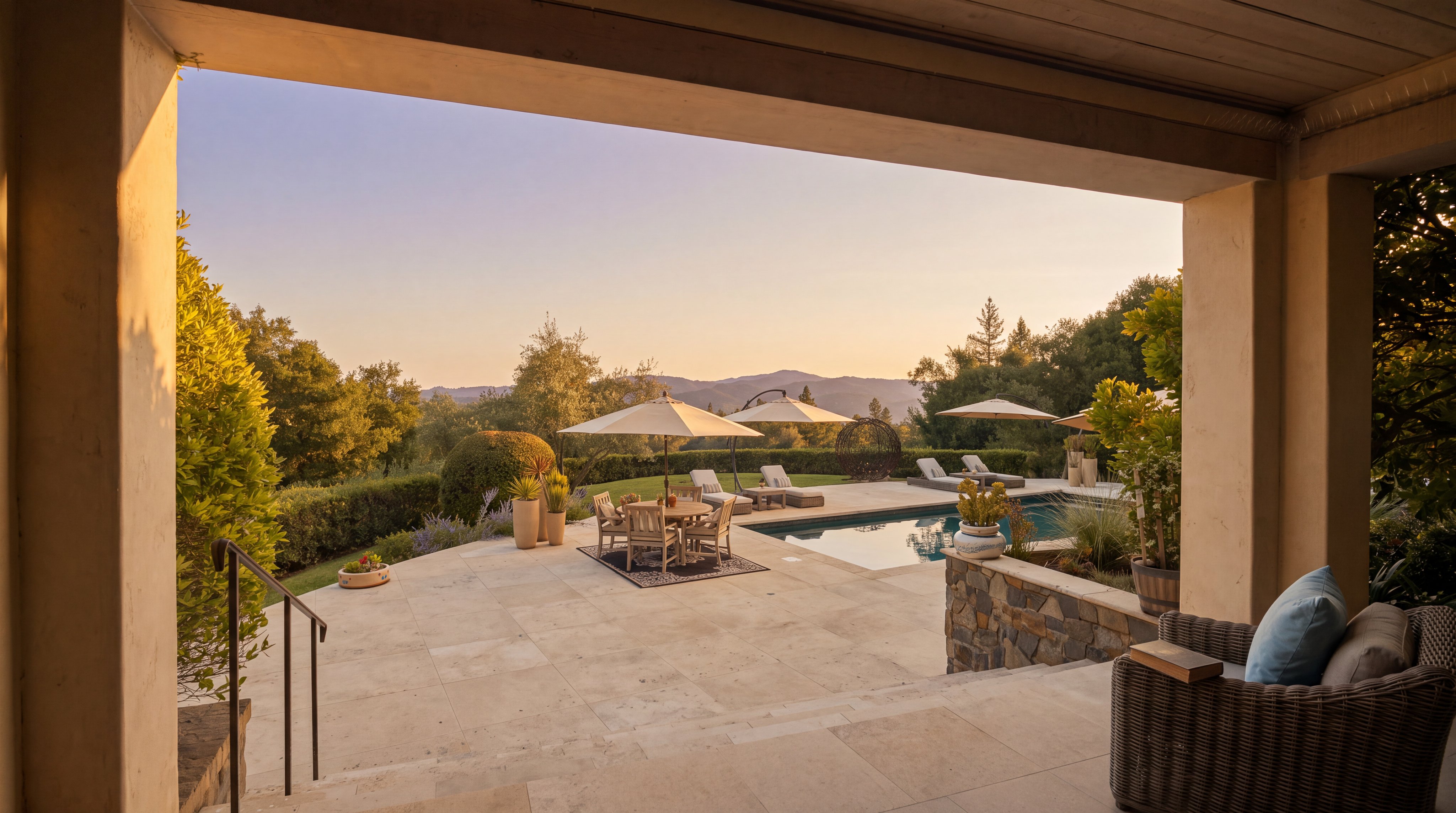 This image captures a spacious, covered stone patio that transitions seamlessly into an expansive outdoor living area featuring a swimming pool and dining space. The scene is framed by the architectural pillars of the covered porch, looking out over a manicured lawn and rolling hills in the distance. The setting is bright and inviting, showcasing a luxurious outdoor lifestyle with comfortable lounge seating and multiple umbrellas for shade.