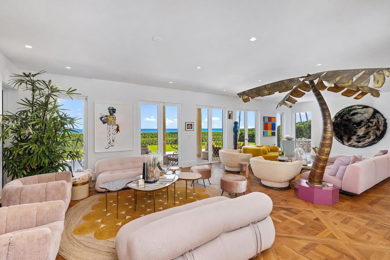 This expansive, light-filled living room features a sophisticated, eclectic design with a prominent sculptural metal palm tree as a focal point. The space is furnished with multiple curved, blush-toned sofas and textured accent chairs arranged on a large, patterned area rug over intricate parquet wood flooring. Large glass doors provide an unobstructed view of the ocean, creating a seamless connection between the luxurious interior and the coastal landscape.