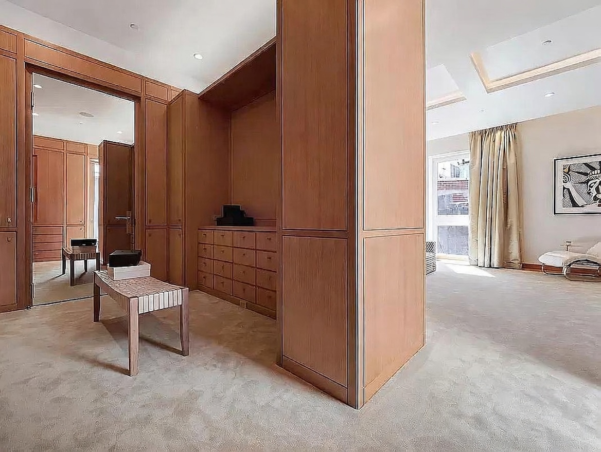 This spacious walk-in closet features extensive light wood cabinetry with numerous drawers and open shelving, providing ample storage. A central wooden bench sits in the foreground, while a large mirror reflects the organized space, leading into a bright, carpeted bedroom area with tall windows and neutral decor. The room exudes a clean, sophisticated, and highly organized aesthetic suitable for a luxury primary suite.