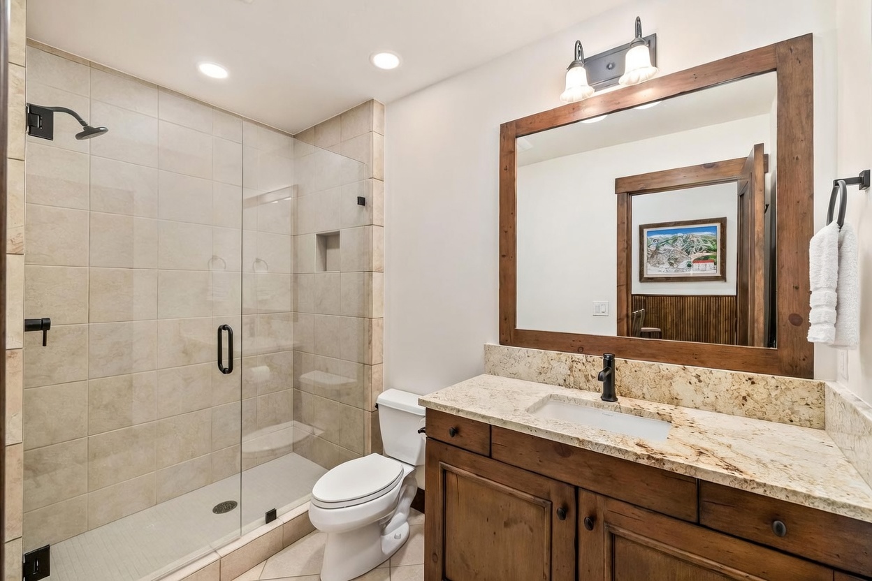 This bathroom features a rustic aesthetic with a wood-framed mirror, a wood vanity, and a walk-in shower enclosed by a glass panel. The shower is finished with neutral-toned wall tiles and includes a built-in niche, while the vanity is topped with a light-colored stone counter. The space is well-lit by recessed ceiling lights and a dual-light fixture above the mirror, creating a warm and functional atmosphere.