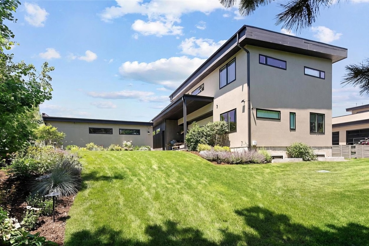 This modern, multi-level home features a contemporary architectural design with clean lines, a flat roof, and a mix of smooth stucco and dark vertical siding. A lush, well-manicured lawn slopes gently toward the house, which includes a covered patio area and several rectangular windows. The scene is captured on a bright, sunny day with a clear blue sky, highlighting the home's sophisticated and minimalist aesthetic.