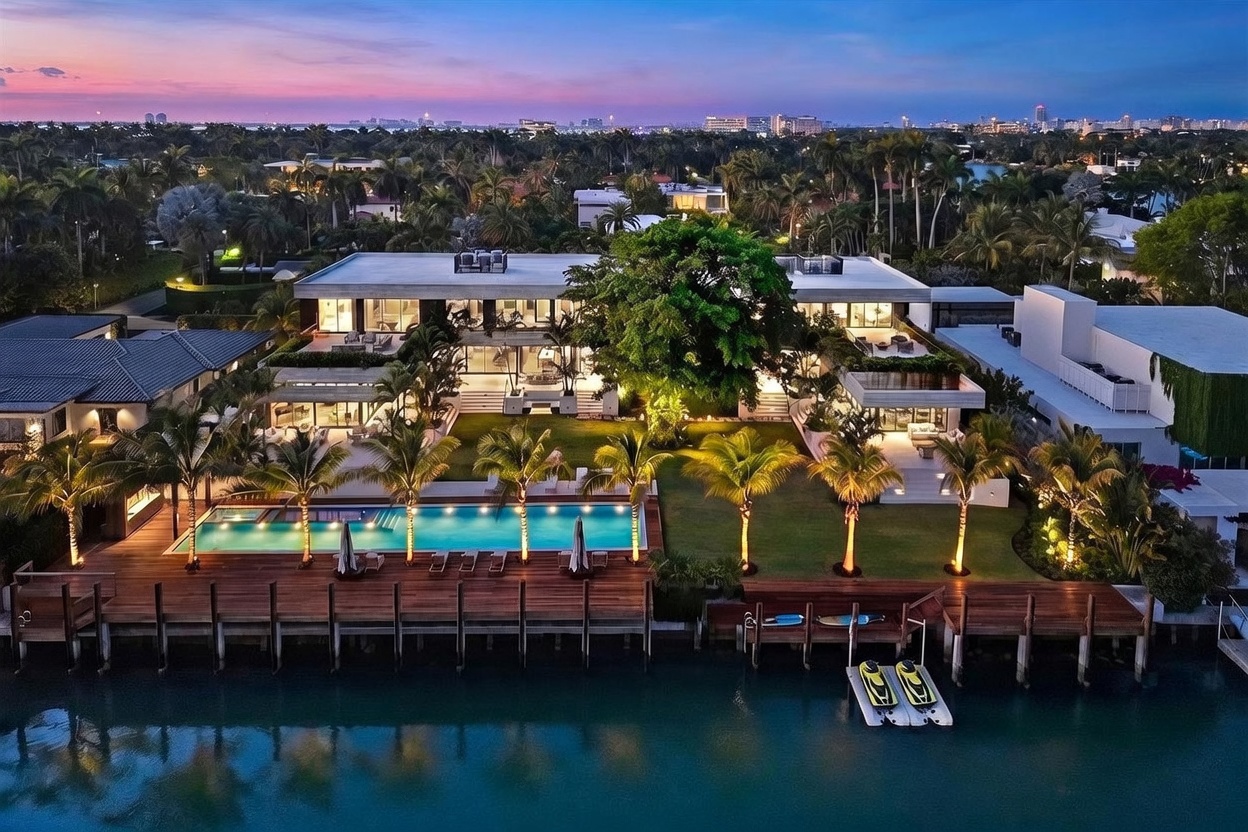 This stunning aerial view captures a luxurious waterfront estate at dusk, showcasing a modern architectural design with expansive glass walls and multiple levels. The property features a pristine swimming pool, a manicured lawn, and a private wooden dock extending into the calm canal, complete with jet skis. The warm interior lighting contrasts beautifully with the twilight sky, creating an inviting and sophisticated atmosphere.
