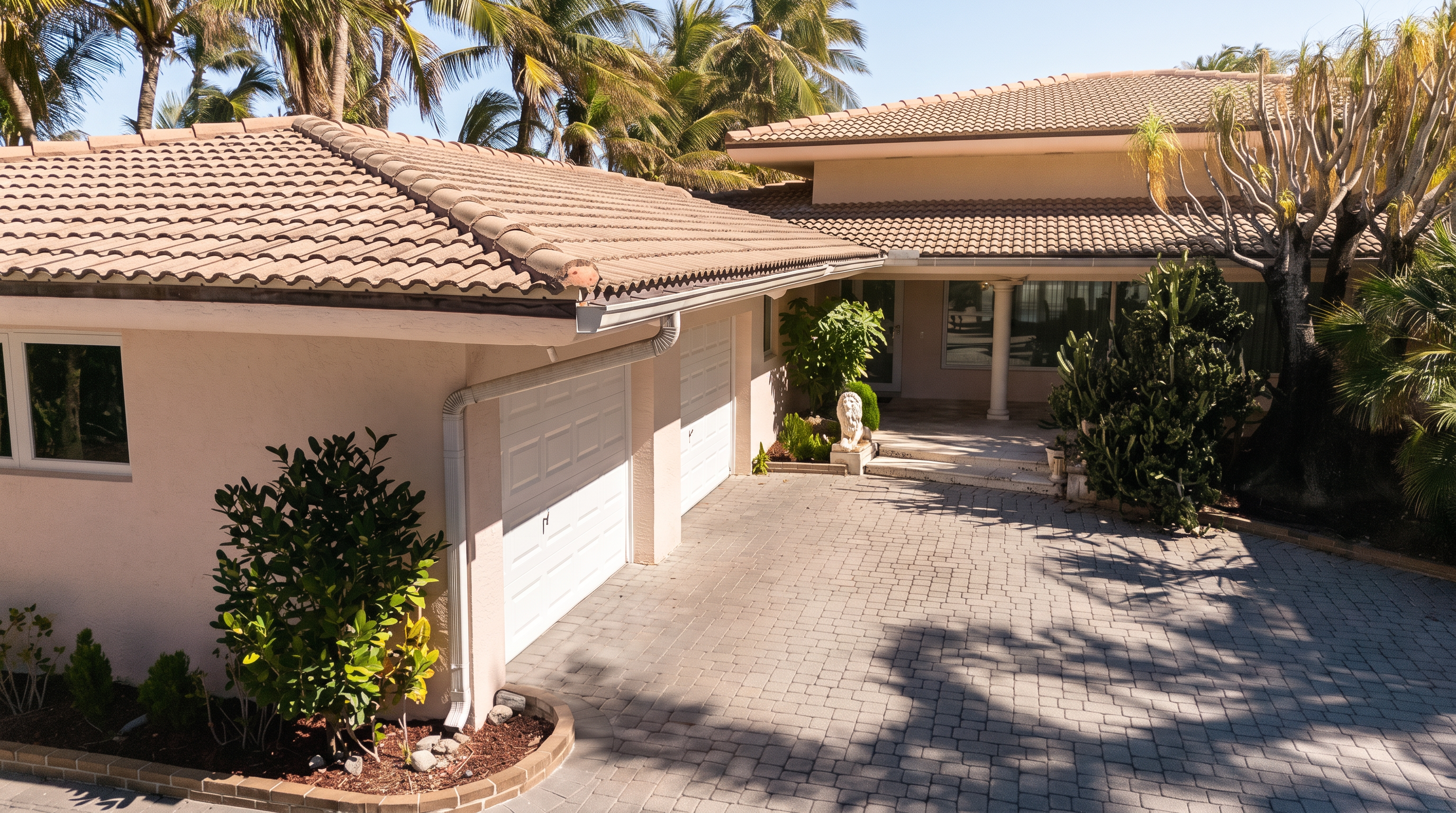 This elevated perspective captures the front exterior of a residence, featuring a multi-car garage with white paneled doors and a paved driveway. The home showcases a light-colored stucco facade, a tiled roof, and a welcoming entryway accented by a decorative lion statue and lush tropical landscaping. The scene is bathed in bright, natural sunlight, highlighting the architectural details and the well-maintained driveway.