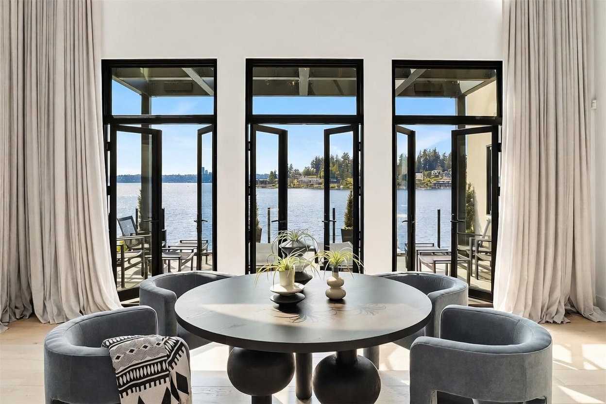 This elegant dining area features a dark, circular table surrounded by four plush, slate-blue upholstered chairs, creating an intimate setting. The space is defined by three sets of tall, black-framed glass doors that provide an expansive, unobstructed view of the lake and surrounding greenery. Long, neutral-toned curtains frame the windows, enhancing the sophisticated and serene atmosphere of this waterfront home.