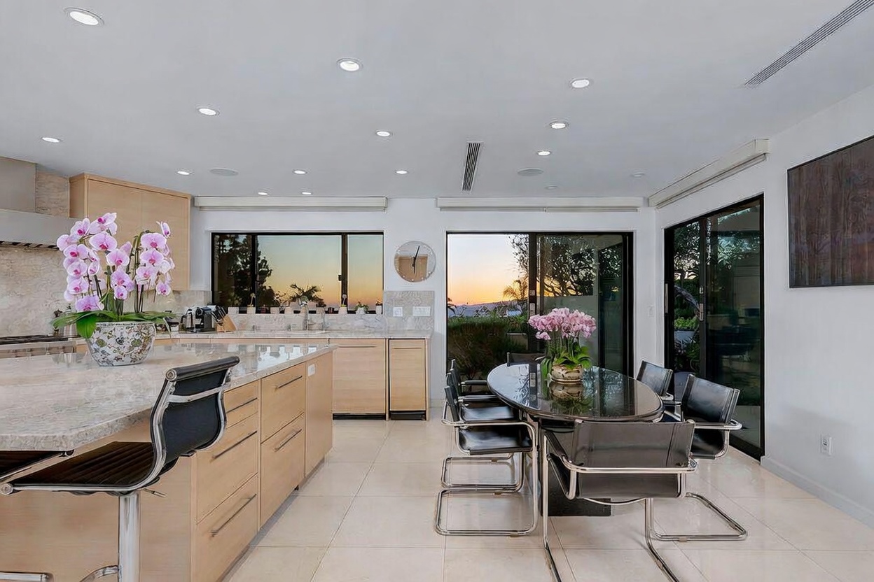 This bright, contemporary kitchen features light wood cabinetry, a large stone-topped island with bar seating, and a dedicated dining area with a dark oval table. Large sliding glass doors provide a seamless transition to the outdoors, allowing natural light to fill the space and offering a view of the sunset. The room is accented by fresh orchids on both the island and dining table, creating an elegant and inviting atmosphere.