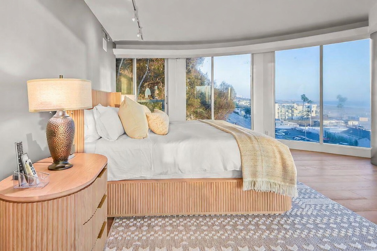 This primary bedroom features a modern, minimalist aesthetic with a light wood bed frame and a matching bedside table. The room is defined by expansive floor-to-ceiling windows that offer a panoramic view of the coastline and beach. A textured, patterned area rug anchors the space, while a warm-toned lamp provides soft lighting against the neutral wall palette.
