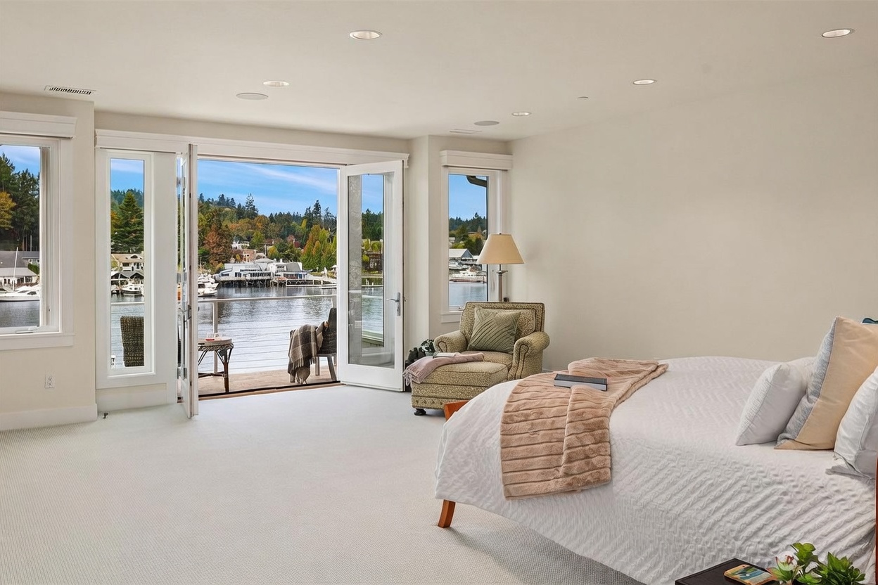 This spacious primary bedroom features neutral carpeting and a large bed dressed in white linens with a soft throw blanket. The room is highlighted by wide glass doors that open onto a private balcony, offering a serene, unobstructed view of the water and surrounding landscape. A comfortable patterned armchair sits near a floor lamp, creating a cozy reading nook that takes full advantage of the natural light and scenic waterfront setting.