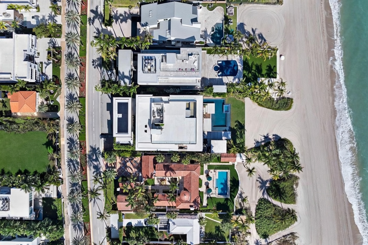 This high-altitude aerial view captures a row of luxury beachfront properties along a coastal road. The scene showcases diverse architectural styles, ranging from modern flat-roofed residences with private pools to more traditional homes with terracotta-tiled roofs, all situated directly against a sandy beach and the ocean. The perspective emphasizes the prime location, lush landscaping, and the seamless transition from private residential estates to the shoreline.