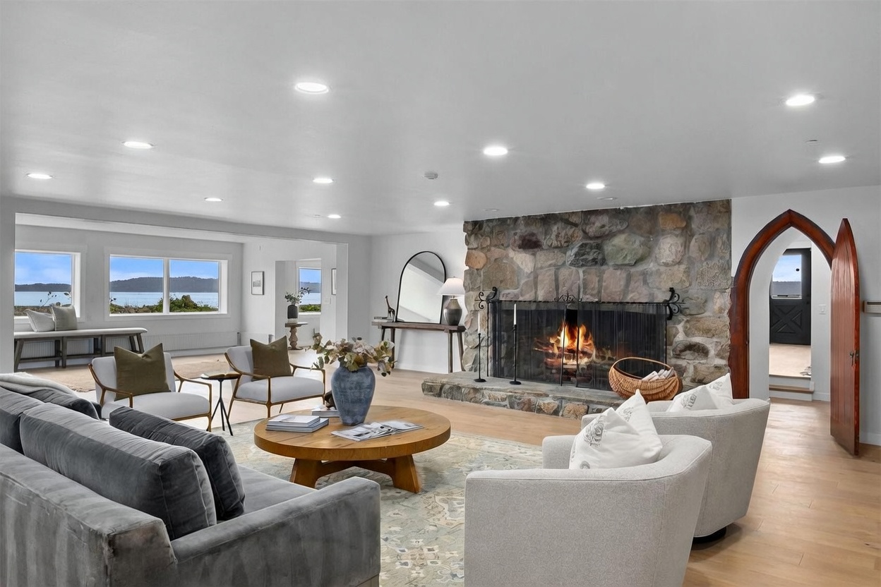 This spacious living room features a prominent stone-clad chimney breast housing a cozy fireplace, serving as the room's focal point. The space is furnished with a plush grey sofa and light-colored armchairs arranged around a large circular wooden coffee table on a patterned area rug. Large windows along the left wall provide expansive views of the water, while an arched wooden doorway adds a unique architectural detail to the right.