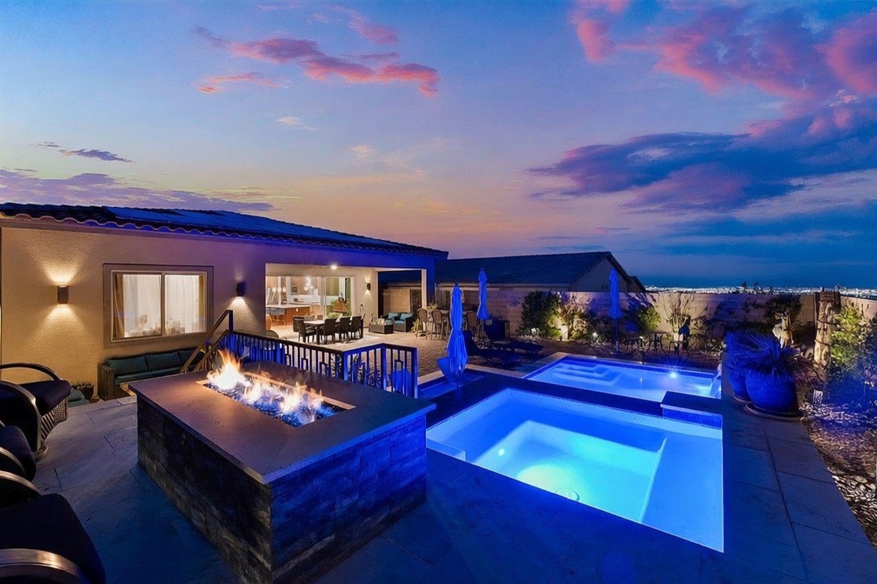 This stunning outdoor living space features a modern rectangular pool and integrated spa, illuminated by soft blue lighting at dusk. A prominent stone-clad fire pit sits on the patio, creating a cozy focal point, while the adjacent covered patio area provides a seamless transition to the home's interior. The scene is set against a dramatic sunset sky, offering a panoramic view of the city lights in the distance.