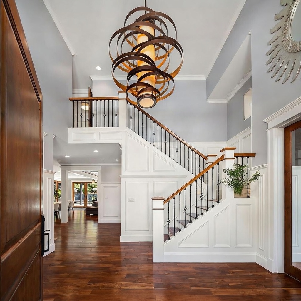 This grand entryway features a dramatic, multi-ringed metal chandelier suspended from a high ceiling above a staircase with dark wood treads and white wainscoting. A console table with decorative vases sits at the base of the stairs, while rich wood flooring extends into the open-concept living space beyond. The perspective is from the front doorway, looking inward to showcase the home's open, airy, and sophisticated architectural design.