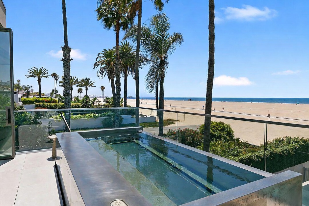 This elevated balcony features a sleek, rectangular plunge pool with a dark, modern finish, positioned to overlook a wide sandy beach and the ocean. A glass railing provides an unobstructed view of the coastal landscape, which is framed by tall palm trees. The space offers a luxurious, serene atmosphere, perfect for enjoying the panoramic seaside scenery.