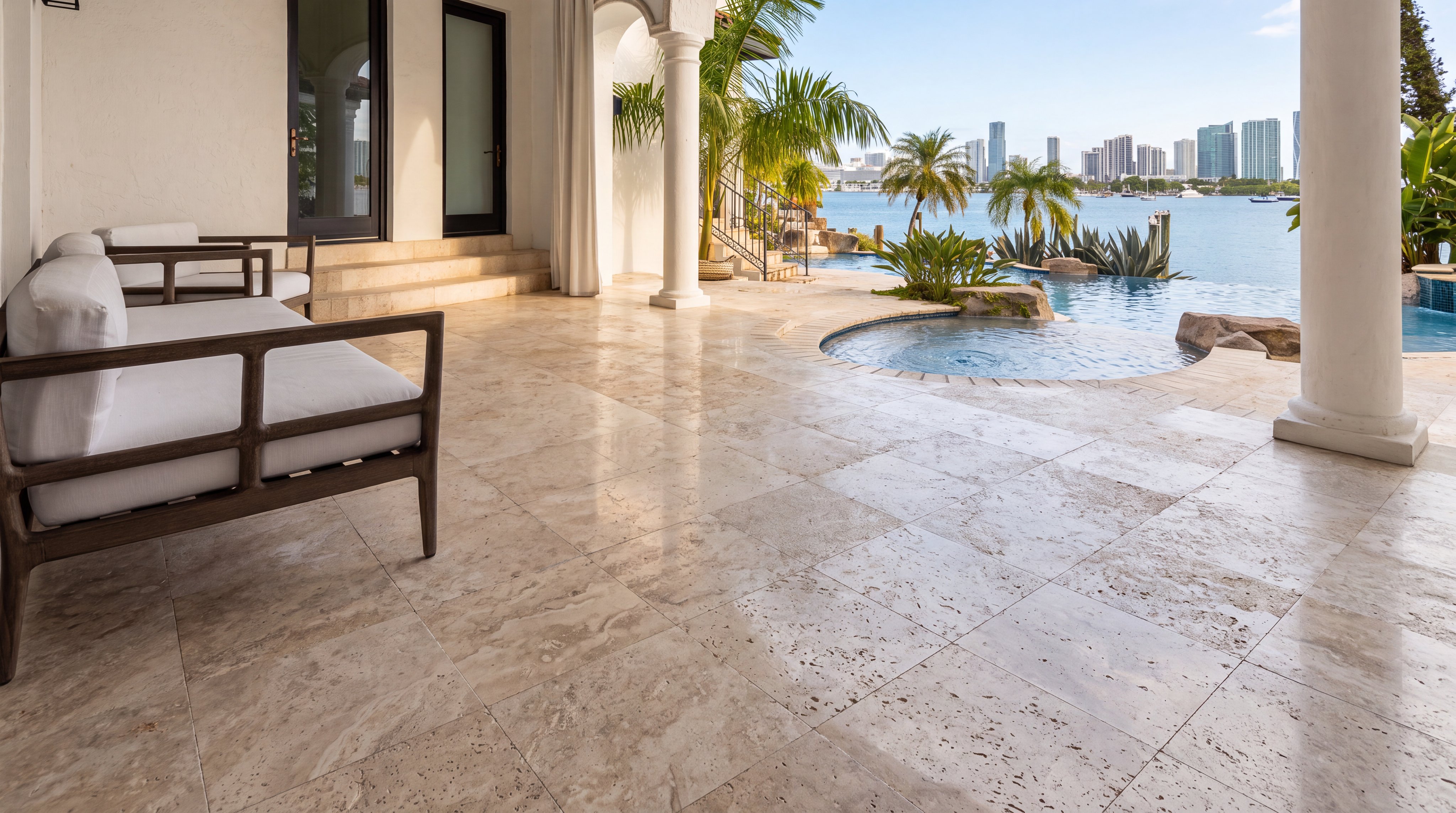 This luxurious outdoor terrace features premium travertine flooring and elegant lounge seating, creating a serene space for relaxation. The area transitions seamlessly into an infinity-edge pool and spa that overlook breathtaking panoramic views of a bay and city skyline. With its columns, lush palm trees, and coastal setting, this space emanates a sophisticated, resort-style ambiance perfect for high-end waterfront living.