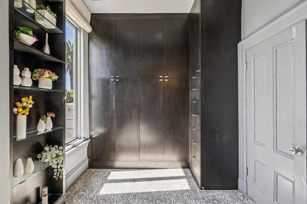 This organized walk-in closet features dark wood cabinetry with integrated drawers and shelving, providing ample storage space. A tall window with a neutral shade allows natural light to illuminate the patterned carpet, while a built-in shelving unit on the left displays decorative items and greenery. The space is accessed through a classic paneled door, creating a functional and sophisticated storage area.