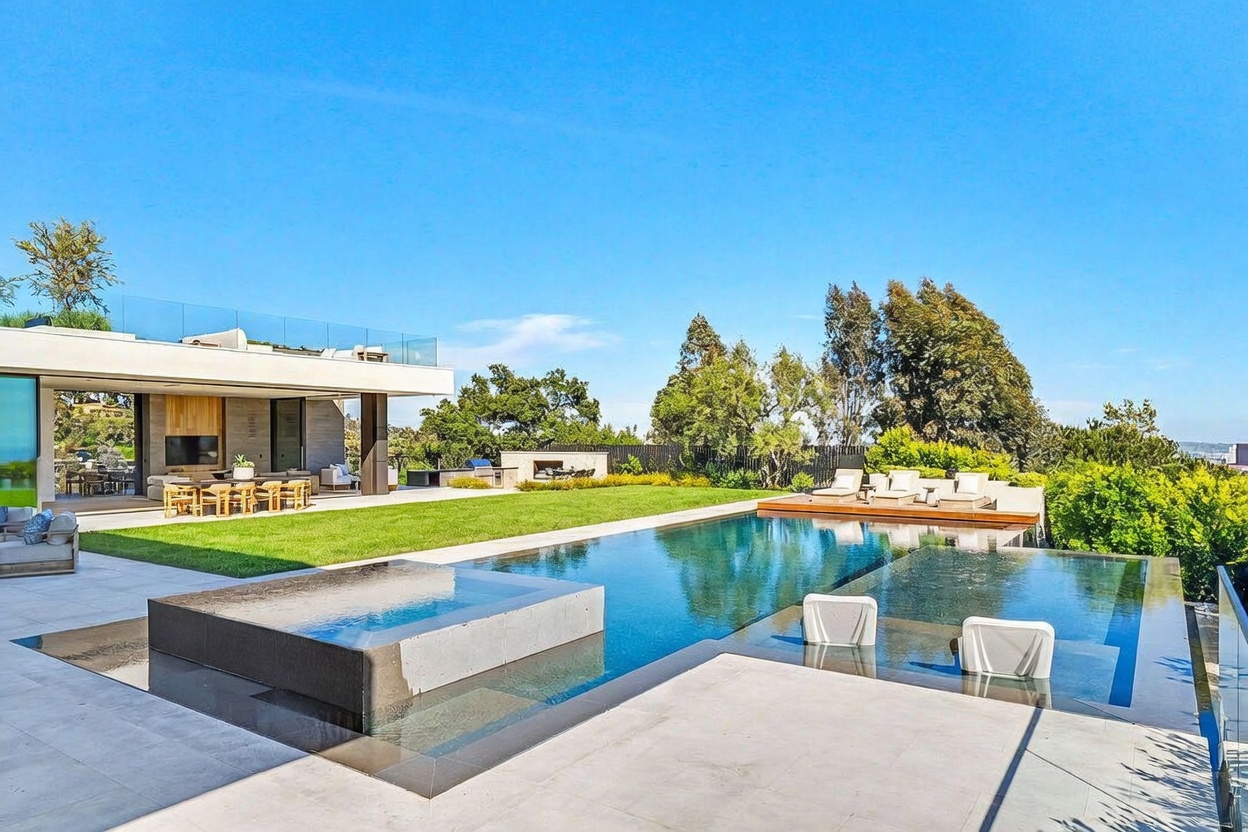 This modern luxury estate features a sprawling backyard with a large infinity-edge pool and an integrated spa. A covered patio area with outdoor seating and a television provides a seamless transition from the indoor living space to the manicured lawn. The scene is captured from a low-angle perspective, emphasizing the clean lines of the architecture and the expansive, sun-drenched outdoor living environment.
