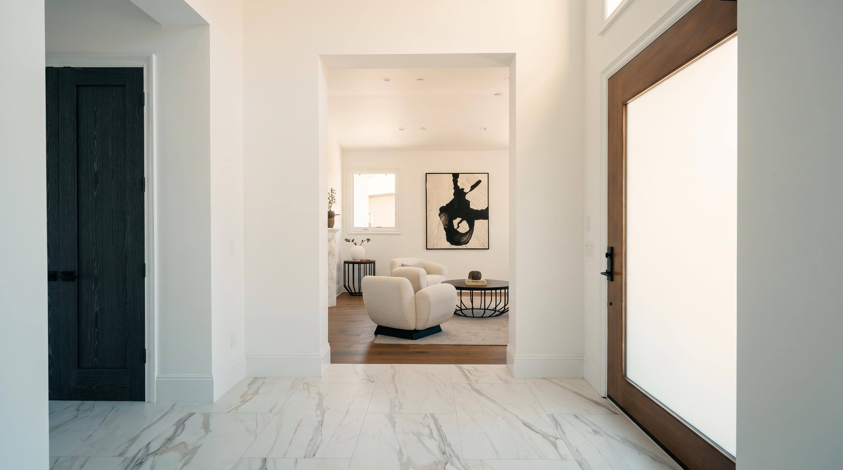 This bright, modern entryway features large-format light-toned stone flooring with subtle veining that leads the eye toward a secondary living space. To the right, a tall, glass-paneled wood door allows natural light to flood the foyer, while a dark, minimalist door stands to the left. The transition into the adjacent room is framed by a clean, white archway, revealing a contemporary seating area with a round coffee table and abstract wall art.