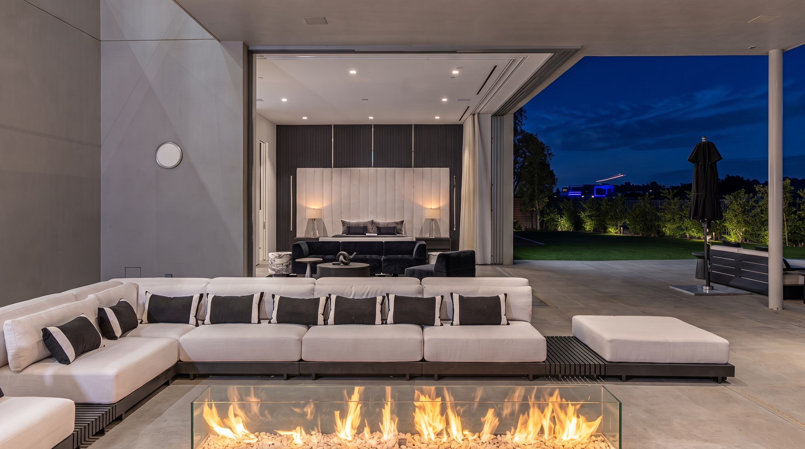 This luxurious covered patio features a large, modern sectional sofa arranged around a rectangular glass-enclosed fire feature. The space seamlessly transitions into an indoor bedroom through wide-opening glass doors, creating an expansive indoor-outdoor living experience. The scene is set at twilight, with soft landscape lighting illuminating the manicured lawn and trees in the background.