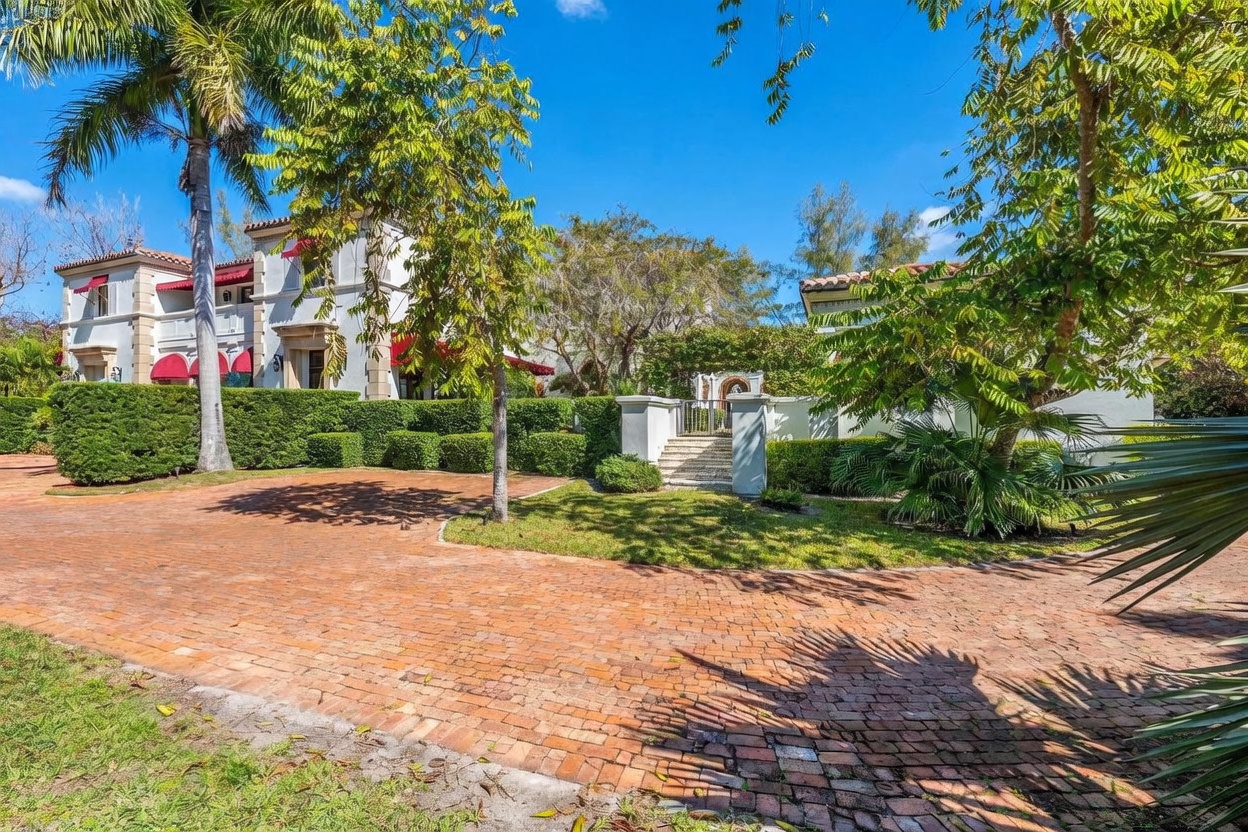 This Mediterranean-style estate features a white stucco facade with red-tiled roofing and prominent red awnings over the windows. A wide, circular brick driveway leads to a landscaped front yard with mature palm trees, manicured hedges, and a stone staircase leading to a gated entrance. The perspective is a wide-angle ground-level shot that captures the grand scale and lush, sunny curb appeal of the property.