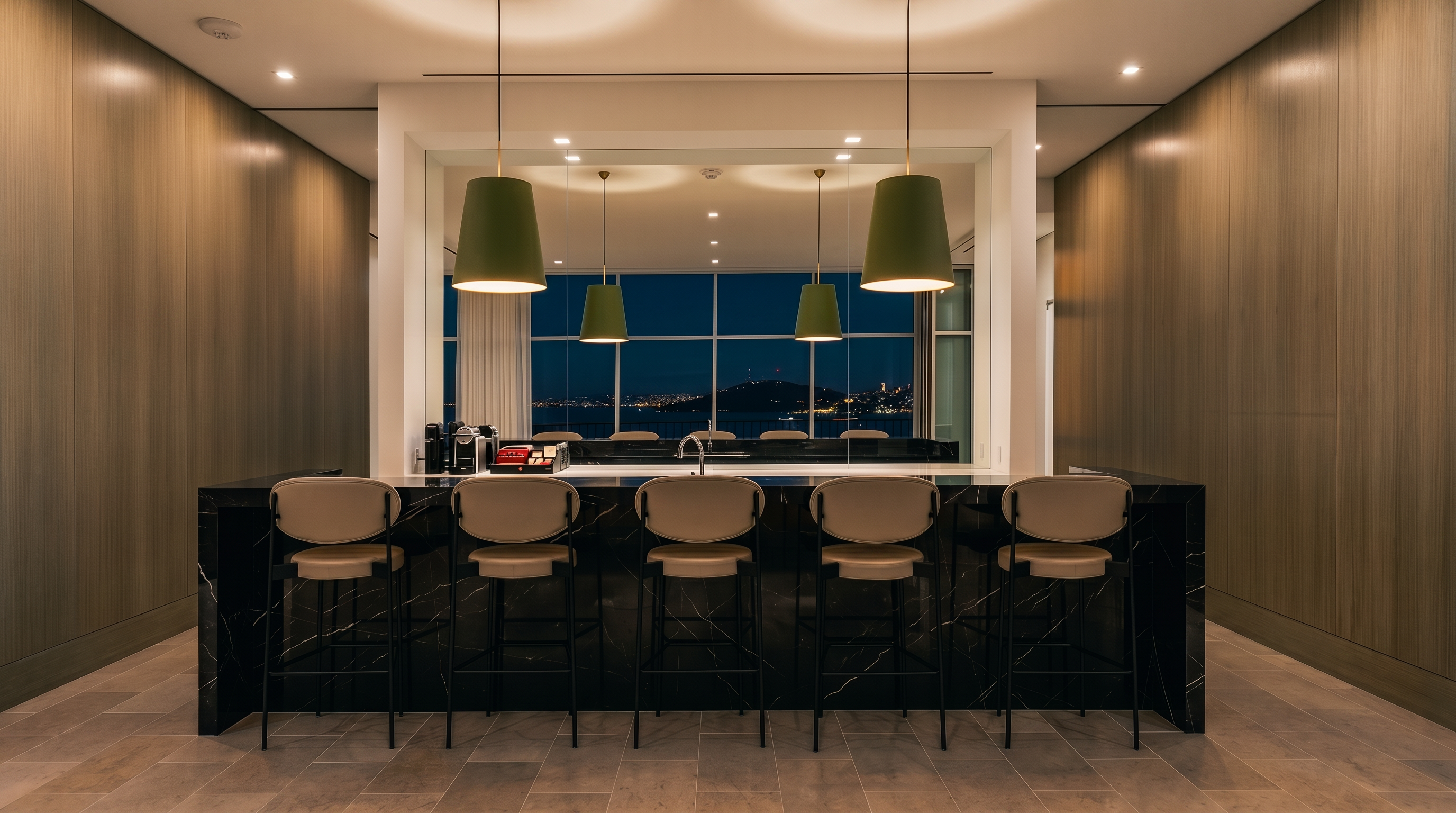 This sophisticated kitchen features a large, dark stone-topped island with five modern bar stools, serving as the central focal point. The space is framed by light wood-paneled walls and illuminated by four distinctive green pendant lights hanging above the island. Large windows in the background provide a clear, expansive view of the water and distant hills, creating a bright and airy atmosphere.