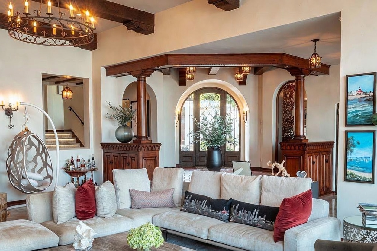 This spacious living room features a large, neutral-toned sectional sofa accented with decorative pillows and a central coffee table. The room is defined by dark wood ceiling beams, a prominent wrought-iron chandelier, and a unique architectural entryway framed by wooden columns and cabinetry. The perspective looks toward a grand arched doorway that leads to an outdoor space, creating an inviting and sophisticated atmosphere.