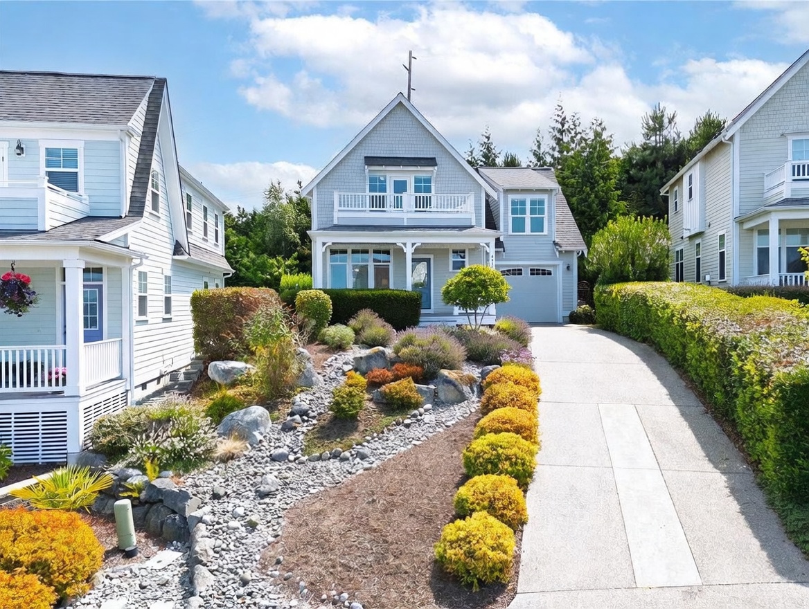 This front-facing view captures a charming, light-blue shingled home with a classic coastal aesthetic. The property features a landscaped front yard with a dry creek bed design, a concrete driveway leading to an attached garage, and a welcoming front porch. The composition highlights the home's architectural symmetry, including a second-story balcony and well-manicured hedges that frame the entrance.