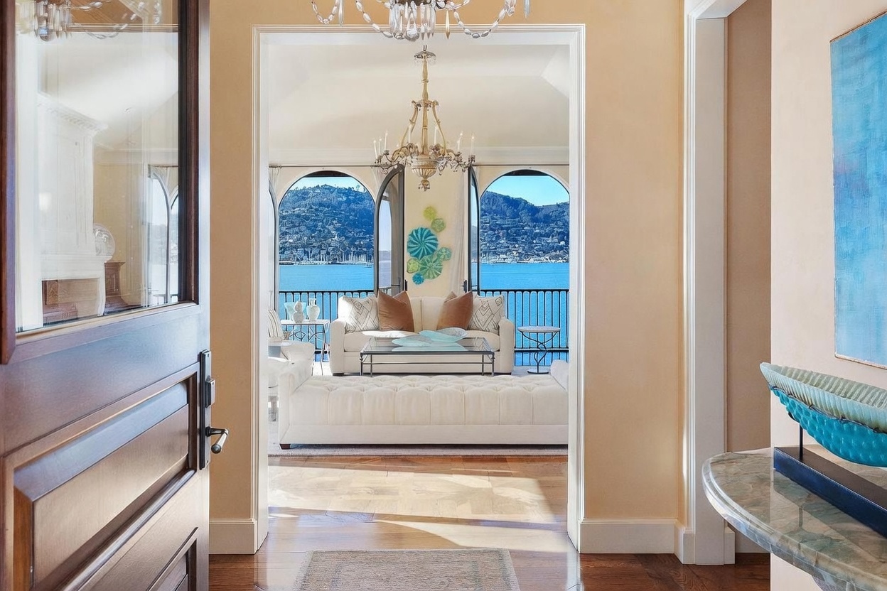 This perspective captures a grand entryway looking through a doorway into a bright, sunlit living room with a stunning waterfront view. The foreground features a polished wood floor and a console table with a decorative glass sculpture, while the living area is furnished with a white sofa, a tufted ottoman, and a glass-topped coffee table. Elegant chandeliers hang in both the hallway and the living space, framing the arched windows that overlook the bay and distant hills.