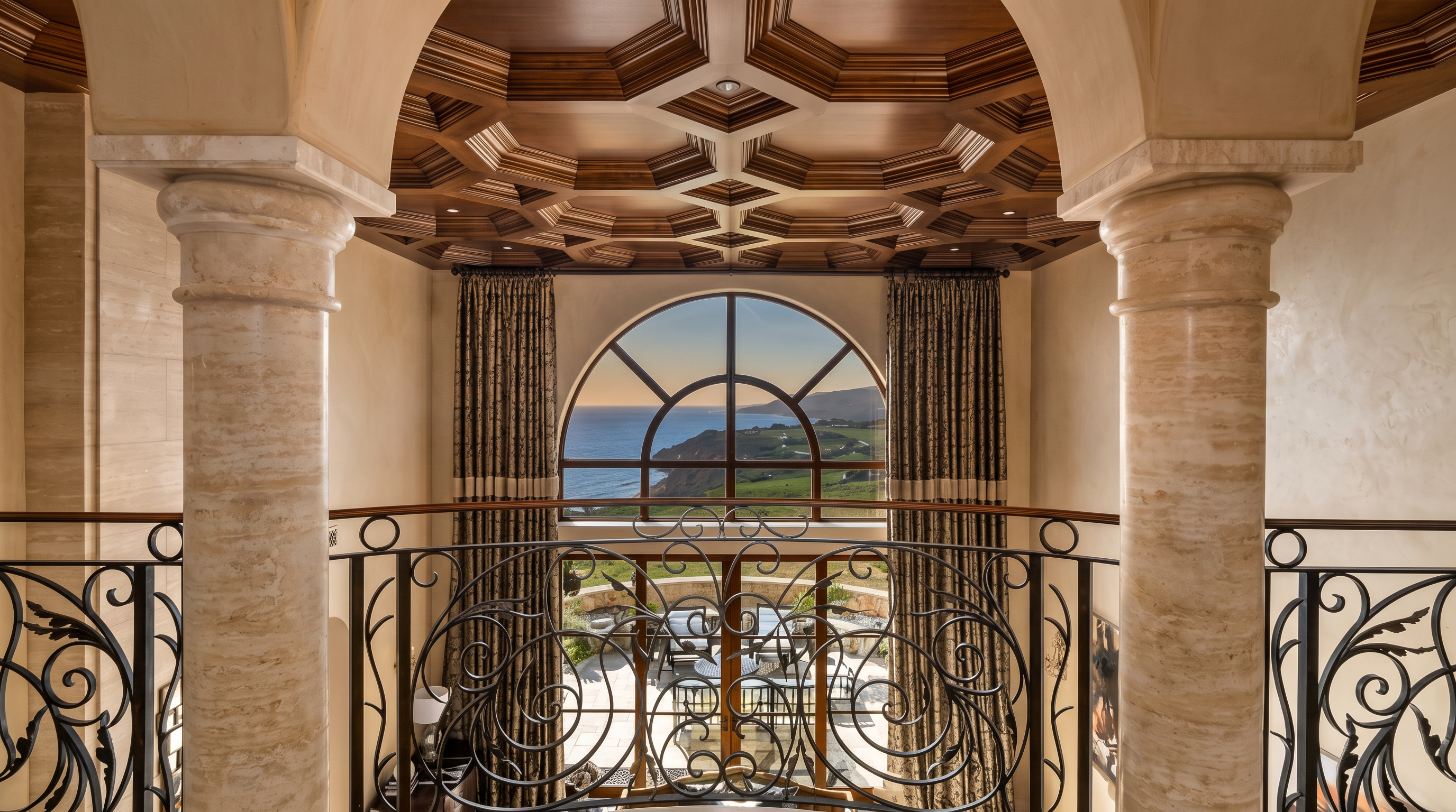 This elegant landing area features a dramatic coffered wood ceiling and two prominent stone columns that frame a large arched window. The view through the window showcases a scenic coastal landscape, while a decorative wrought iron railing lines the balcony. The space exudes a sophisticated, Mediterranean-inspired aesthetic with warm, neutral tones.