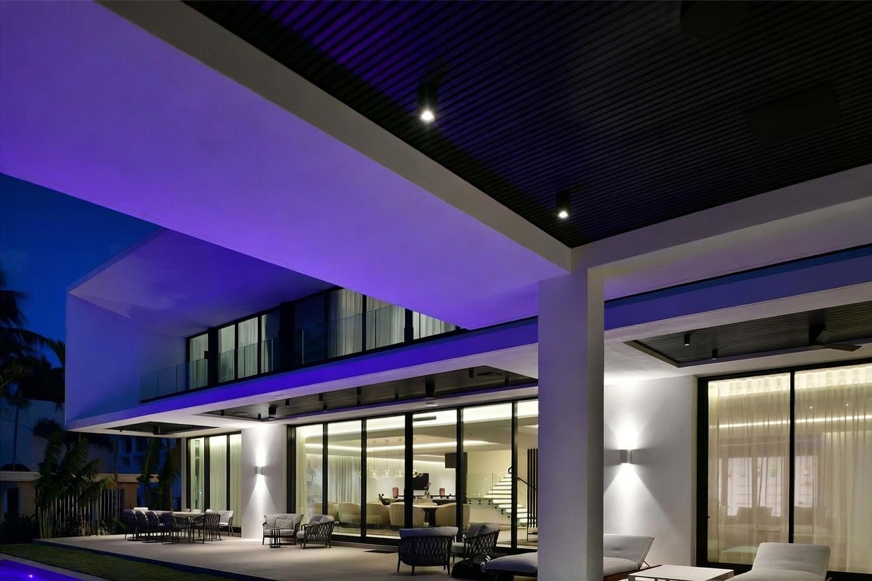This image captures a luxurious, modern outdoor living space at dusk, featuring a spacious covered patio with recessed lighting and a vibrant purple accent glow. The architecture showcases clean lines, large glass sliding doors that reveal a glimpse of the interior, and a contemporary aesthetic. The scene is framed from a low-angle perspective, emphasizing the expansive patio area and the sophisticated transition between the indoor and outdoor environments.