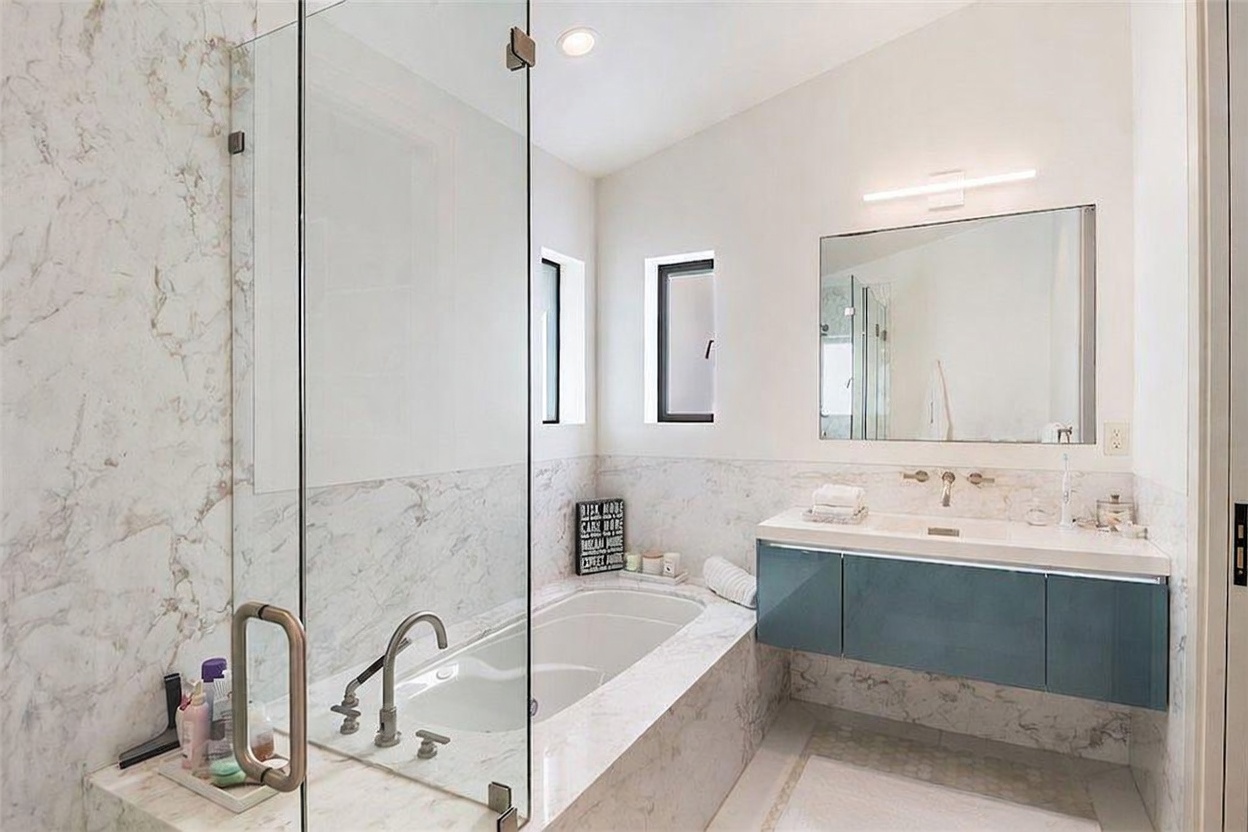 This primary bathroom features a sleek, modern aesthetic with a large soaking tub integrated into a stone-clad surround that matches the wall tiling. A floating vanity with a dark blue finish and a white countertop sits beneath a large, frameless mirror, while a glass-enclosed shower area adds to the open feel. The space is illuminated by a minimalist light fixture and two narrow vertical windows, creating a bright and sophisticated atmosphere.