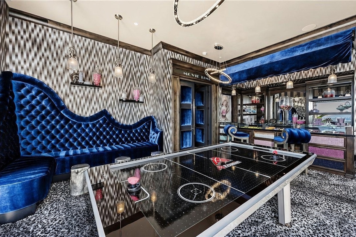 This vibrant, high-energy game room features a sleek black air hockey table as the central focal point, set against a backdrop of bold, patterned wallpaper and a plush, tufted blue velvet sofa. The space includes a built-in bar area with decorative shelving and a doorway marked 'MOVIE TIME,' suggesting a dedicated entertainment zone. The room is accented by modern, circular light fixtures and a unique, textured carpet, creating a luxurious and playful atmosphere.